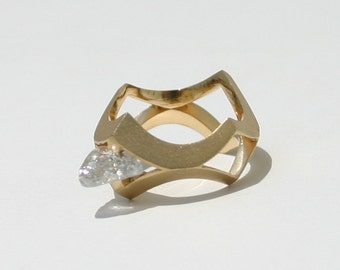May include: A gold ring with a unique design featuring a single, clear diamond set in the center. The ring has a modern, geometric shape.