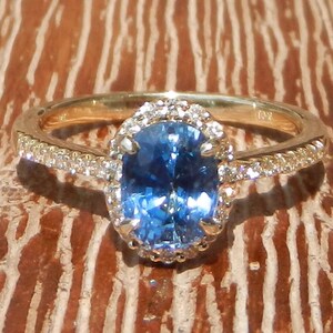 May include: A gold ring with a blue oval gemstone surrounded by a halo of smaller diamonds. The ring has a delicate band with a row of diamonds.