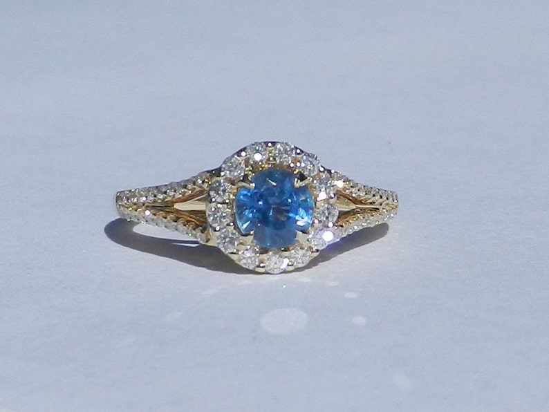 Op de afbeelding: Een gouden ring met een blauwe edelsteen omringd door een halo van kleinere diamanten. De ring heeft een gespleten band met een gevlochten ontwerp.