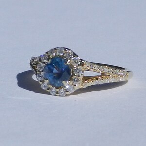 Op de afbeelding: Een gouden ring met een blauwe edelsteen omringd door een halo van kleinere diamanten. De ring heeft een gespleten schacht met diamanten die langs de band zijn gezet.