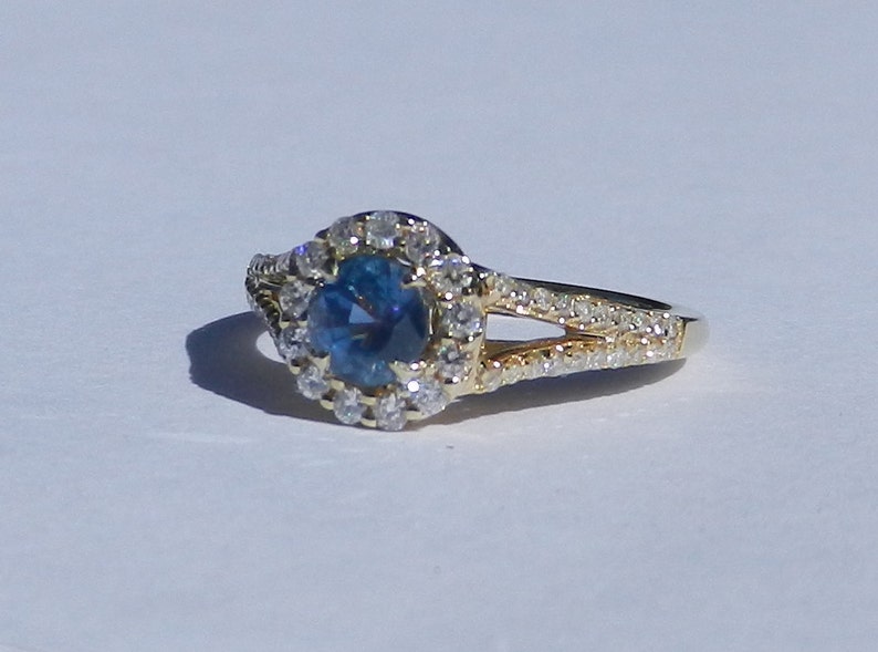 Op de afbeelding: Een gouden ring met een blauwe edelsteen omringd door een halo van kleinere diamanten. De ring heeft een gespleten schacht met diamanten die langs de band zijn gezet.