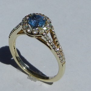 Op de afbeelding: Een gouden ring met een blauwe edelsteen omringd door een halo van kleinere diamanten. De ring heeft een gespleten schacht met diamanten die langs de band zijn gezet.