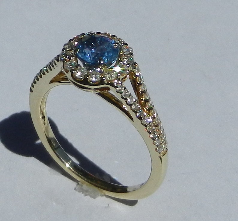 Op de afbeelding: Een gouden ring met een blauwe edelsteen omringd door een halo van kleinere diamanten. De ring heeft een gespleten schacht met diamanten die langs de band zijn gezet.