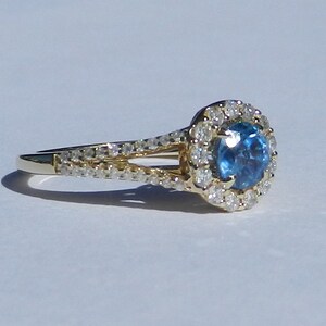 Op de afbeelding: Een gouden verlovingsring met een blauwe edelsteen omringd door een halo van kleinere diamanten. De ring heeft een gesplitste schacht met diamanten die langs de band zijn gezet.
