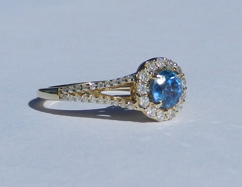 Op de afbeelding: Een gouden verlovingsring met een blauwe edelsteen omringd door een halo van kleinere diamanten. De ring heeft een gesplitste schacht met diamanten die langs de band zijn gezet.
