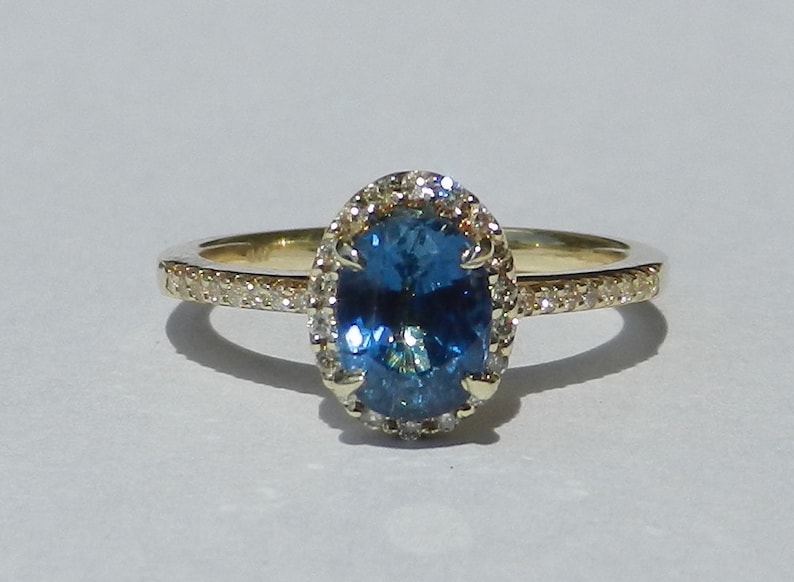 May include: A gold ring with a blue oval gemstone surrounded by a halo of smaller diamonds. The band is also set with smaller diamonds.