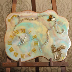 Pode incluir: Um relógio de parede decorativo com uma forma orgânica única. O mostrador do relógio apresenta algarismos romanos dourados sobre um fundo pastel com um design em espiral. Adornado com detalhes dourados e elementos decorativos.