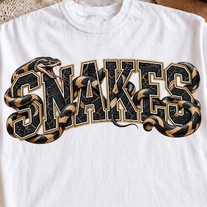 Può includere: Una t-shirt bianca a maniche corte con una grande stampa grafica della parola SNAKES in grassetto nero con contorni dorati. Un lungo serpente sinuoso con squame nere e dorate è avvolto attorno e attraverso le lettere.