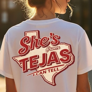 Puede incluir: Camiseta blanca con un gráfico rojo y blanco en la espalda que dice "She's from TEJAS I CAN TELL" en una fuente estilizada y un contorno de Texas. La camiseta la lleva una persona con el pelo recogido en un moño.