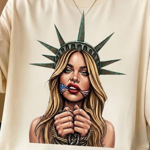 May include: Cream-colored t-shirt featuring a graphic of a woman with long blonde hair, wearing the Statue of Liberty crown, and an American flag in her mouth. The woman's hands are bound with rope. The image has a distressed, artistic style.