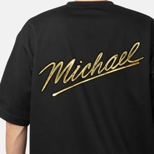 May include: Black t-shirt with the name "Michael" in a gold, cursive font across the back. The text has a 3D effect, giving it a raised appearance. The shirt has short sleeves and a crew neck.