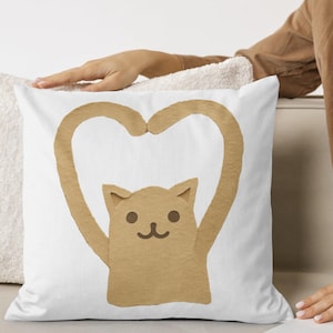 May include: White square throw pillow featuring a tan cat design forming a heart shape with its arms. The cat has a simple, smiling face. The pillow is on a white surface.