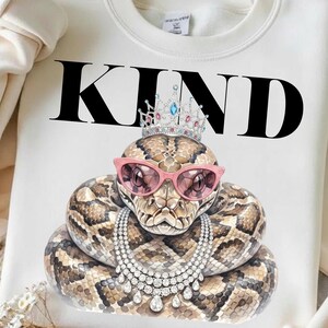 Könnte beinhalten: Weißes Sweatshirt mit dem Wort "KIND" in schwarzen Blockbuchstaben. Eine Schlange mit rosa Sonnenbrille, einer Juwelenkrone und einer Perlenkette ist darunter zentriert. Die Schlange ist braun und beige mit einem detaillierten Muster.