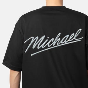 Puede incluir: Camiseta negra con el nombre "Michael" en cursiva, con un efecto de pedrería plateada en la espalda. El texto es plateado y la camiseta tiene mangas cortas.