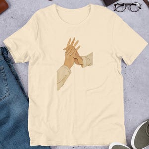 May include: Cream-colored short-sleeve t-shirt featuring an illustration of two hands clasped together. The graphic is in a minimalist style, with the hands depicted in shades of brown and beige. The shirt is paired with blue jeans and gray sneakers.