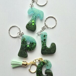 May include: Four resin letter keychains in the shapes of T, P, L, and G. Each letter features a gradient colour transition from light mint green at the top to a deep forest green at the base, embedded with sparkling green glitter throughout.