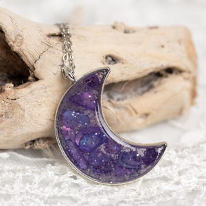 May include: A silver-coloured chain necklace featuring a crescent moon pendant. The moon pendant is filled with a swirling mix of purple, blue, and silver glitter, set in a silver-toned bezel. The necklace is displayed on driftwood.