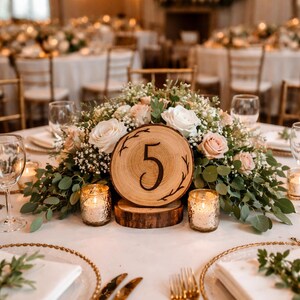 May include: A wedding reception table setting with a floral centerpiece featuring white and pink roses, greenery, and a wooden table number. Gold-rimmed plates, silverware, and lit candles add to the elegant decor. Overhead, string lights create a warm ambiance.