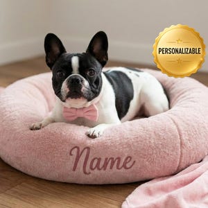 以下が含まれることがあります： ピンクの蝶ネクタイをした白黒のフレンチブルドッグが、「Name」の刺繍が入ったピンクの犬用ベッドで休んでいます。ベッドはピンクの毛布に囲まれています。「Personalizable」と書かれた金のバッジが背景にあります。