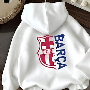 Könnte beinhalten: Weißer Kapuzenpullover mit dem FC Barcelona-Logo in Rot und Blau. Das Logo zeigt ein Schild mit Kreuz und Streifen sowie den Text "FCB" und "BARÇA".