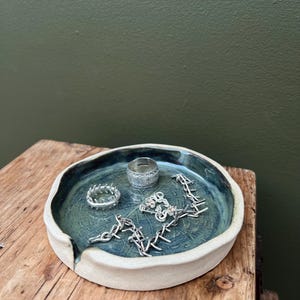 May include: A handmade ceramic jewelry dish with a teal interior and a white rim. The dish holds a silver ring, a silver chain, and another silver ring. The dish sits on a wooden surface.