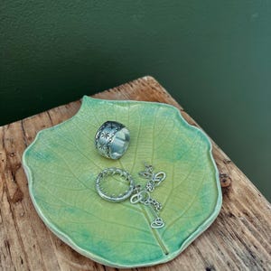 Puede incluir: Un plato de cerámica verde claro en forma de hoja contiene anillos y un collar de plata. El plato tiene una superficie texturizada y un degradado de verde y azul. Los anillos incluyen una banda ancha con diseños de estrellas y una banda fina y texturizada.