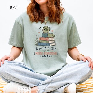 Könnte beinhalten: Ein salbeigrünes T-Shirt mit einer Grafik eines Zeichentrickwurms, der eine Brille trägt und auf einem Stapel aus drei Büchern liest. Der Text lautet A BOOK A DAY keeps the MENTAL BREAKDOWN AWAY, ergänzt durch kleine Herz- und Sternmotive.