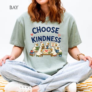 May include: A sage green t-shirt featuring a graphic of six cartoon ducks sitting around a table. The text CHOOSE KINDNESS is printed in dark blue above the ducks. One duck holds a sign that reads EVERYONE BELONGS HERE. The design includes books.