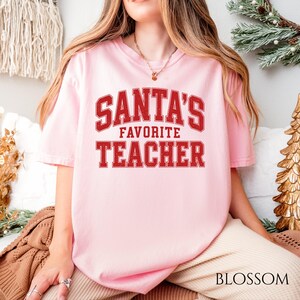 Può includere: Una t-shirt a maniche corte rosa chiaro con il testo rosso SANTA'S FAVORITE TEACHER in un carattere in stile college. La maglietta ha un girocollo e una vestibilità rilassata, con la grafica centrata sul petto in una texture dall'aspetto usurato.