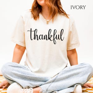 Puede incluir: Una camiseta de manga corta color marfil con la palabra thankful impresa en letra cursiva negra en el pecho. La camiseta tiene cuello redondo y se combina con unos vaqueros azul claro. El texto IVORY es visible en la esquina superior derecha.