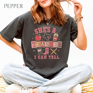 Peut inclure: Un t-shirt gris anthracite à manches courtes avec une impression graphique portant le texte "SHE'S A TEACHER I CAN TELL". Le motif comprend une pomme rouge, le contour de l'État du Texas, un gobelet à café, un carnet, une botte de cow-boy rose et deux crayons.
