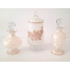 May include: Three vintage glass perfume bottles and jars. Two bottles have a textured, pearlescent finish and clear glass stoppers. The central jar is white with a floral design and a clear glass lid.