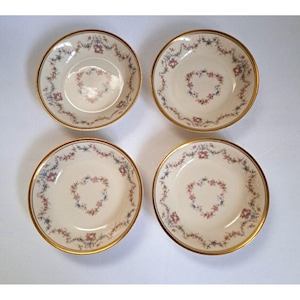 May include: A set of four round ceramic bowls with a cream-colored base, featuring a delicate floral garland pattern in pink and blue around the rim and a central heart-shaped floral motif, finished with a thin gold-toned trim on the edges.
