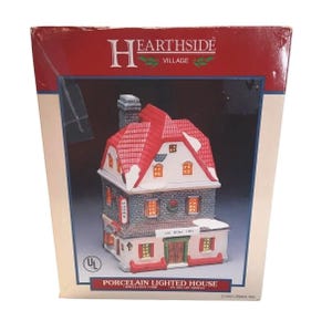 May include: A Hearthside Village porcelain lighted house in its original box. The miniature building is the Ox Bow Inn, featuring a red roof with snow, a stone-textured middle floor, and a white ground floor with a green door and lit windows.