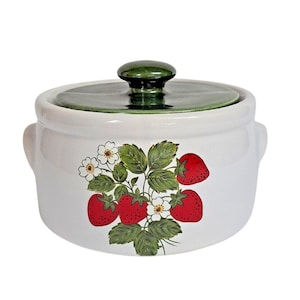 May include: A white ceramic lidded casserole dish featuring a painted design of red strawberries, green leaves, and white flowers on the front. The lid is a solid dark green colour with a central knob handle and two side handles on the main body.