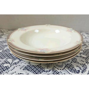 May include: A stack of three round, cream-colored soup bowls with wide rims. Each bowl features a delicate floral pattern in soft pink, blue, and green hues, accented by a thin brown band and a gold-toned trim along the outer edge.