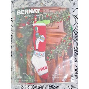 以下が含まれることがあります： 透明なプラスチックパッケージに入ったBernatのクリスマスストッキング編み物キット。ストッキングは赤と白で、ドラマーのグラフィックとPaulという名前が入っています。パッケージには暖炉のシーン、Drummerという文字、48cmというサイズが記載されています。