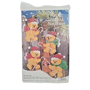 May include: A plastic canvas ornament kit in a clear plastic bag featuring four yellow teddy bears wearing red Santa hats and scarves. The packaging displays the text PLASTIC CANVAS ORNAMENT KIT and states the kit makes four 4 inch ornaments.