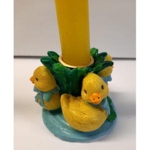 May include: A yellow taper candle sits in a circular candle holder featuring three yellow ducklings with orange beaks and blue bows. The base is painted light blue, and the central holder is surrounded by green leaves, creating a decorative piece.