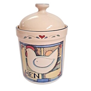 May include: A cream-colored ceramic canister with a matching lid, featuring a hand-painted illustration of a white hen, a sunburst, and the word HEN in black lettering. The design includes a small red heart and green leaf accents near the rim.