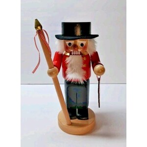May include: A wooden nutcracker figurine with a red jacket, black pants, and a black top hat. The figurine has a white beard and holds a staff with a red and white ribbon. It stands on a round wooden base.