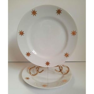 May include: Two white ceramic plates featuring a repeating pattern of metallic gold eight-pointed stars around the rim. The plates are displayed on a gold-toned metal stand, with one plate positioned vertically and the other resting horizontally below.