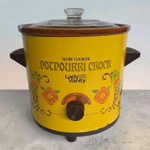 Puede incluir: Una olla de cocción lenta amarilla Lady Vanity Potpourri Crock con tapa de cristal transparente. El frente presenta texto marrón, un gráfico floral y una perilla de control circular con ajustes de bajo y alto. Tiene asas laterales y base marrón.