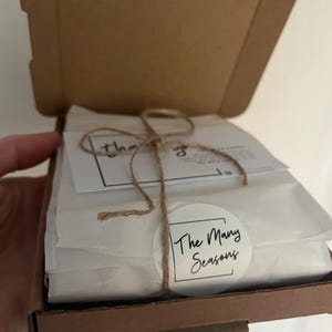 May include: A brown cardboard shipping box containing white tissue paper wrapped items tied with thin brown twine. A circular white sticker on the front features the black text The Many Seasons inside a square border, with a thank you note tucked in.