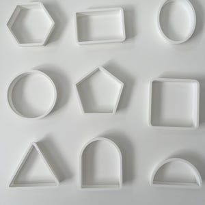 May include: A collection of nine white plastic geometric cookie cutters arranged in a grid. The shapes include a hexagon, rectangle, oval, circle, pentagon, square, triangle, arch, and semicircle, all featuring clean, open-frame designs.