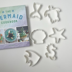 May include: A cookbook titled The Mermaid Cookbook by Alix Carey with a pastel marbled cover, accompanied by six white plastic cookie cutters in the shapes of a mermaid tail, sea turtle, seashell, starfish, seahorse, and a tropical fish.