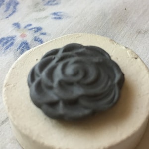May include: A small, round, white ceramic base with a black, raised, rose design on top.