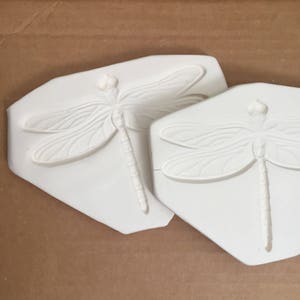 May include: Two white plaster molds of dragonflies. The molds are detailed and show the wings, body, and legs of the dragonfly.