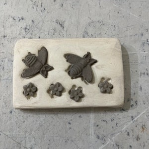 May include: A white ceramic mold with two bee and five flower impressions. The mold is designed for making polymer clay or resin crafts.