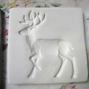 May include: White ceramic tile with a raised design of a deer with antlers. The deer is facing left and has a detailed, textured body.